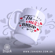 Чашка Ти – мій вибір, біла з яскравим принтом, показана з легким нахилом на світло-сірому фоні, з написом "Ти – мій вибір" та сердечками, має гладку ручку та рівний край, з глянцевою поверхнею.