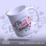 Чашка Ти – мій вибір, біла з яскравим принтом, показана з легким нахилом на світло-сірому фоні, з написом "Ти – мій вибір" в чорному і червоному кольорах, має гладку ручку та рівний край, з глянцевою поверхнею.