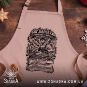 Фартух Бог барбекю, бежевого кольору, зображений з верхнього ракурсу на дерев'яному фоні, принт з малюнком барбекю та текстом на чорному фоні.