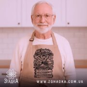 Фартух Бог барбекю, бежевого кольору, зображений з верхнього ракурсу на дерев'яному фоні, принт з малюнком барбекю та текстом на чорному фоні.