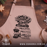 Фартух майстер шампура, бежевого кольору, зображений з верхнього ракурсу на дерев'яному фоні, має чорний принт з текстом та графікою, поруч розташовані святкові елементи.