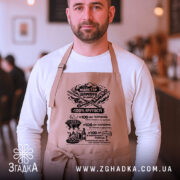 Фартух майстер шампура, бежевого кольору, зображений на чоловікові з переднього ракурсу в кафе, має чорний принт з текстом та графікою, чоловік позує з серйозним виразом обличчя.