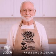 Фартух майстер шампура, бежевого кольору, зображений з верхнього ракурсу на дерев'яному фоні, має чорний принт з текстом та графікою, поруч розташовані святкові елементи.