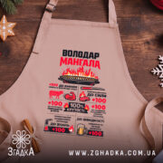 Фартух володар мангала бежевого кольору, зображений з близького ракурсу на дерев'яному фоні, принт яскравий, з текстом і малюнками, що підкреслюють кулінарні навички.