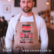 Фартух володар мангала бежевого кольору, зображений на чоловікові в кафе, фон розмитий, принт яскравий, з текстом і графікою, що стосуються кулінарії.