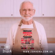 Фартух володар мангала бежевого кольору, зображений з близького ракурсу на дерев'яному фоні, принт яскравий, з текстом і малюнками, що підкреслюють кулінарні навички.