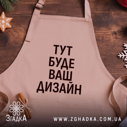 Фартух з вашим дизайном, бежевого кольору, зображений з верхнього ракурсу на дерев'яному фоні, навколо декор з новорічними елементами, принт чорного кольору з текстом 'ТУТ БУДЕ ВАШ ДИЗАЙН'.
