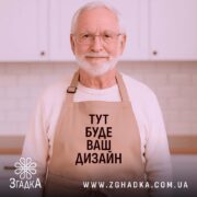 Фартух з вашим дизайном, бежевого кольору, зображений з верхнього ракурсу на дерев'яному фоні, навколо декор з новорічними елементами, принт чорного кольору з текстом 'ТУТ БУДЕ ВАШ ДИЗАЙН'.