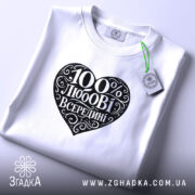 Футболка 100% любові всередині, біла, складена на поверхні, ракурс зверху, фон світло-фіолетовий, принт у формі серця чорного кольору з написом "100% любові всередині" в декоративному шрифті, з етикеткою зеленого кольору.