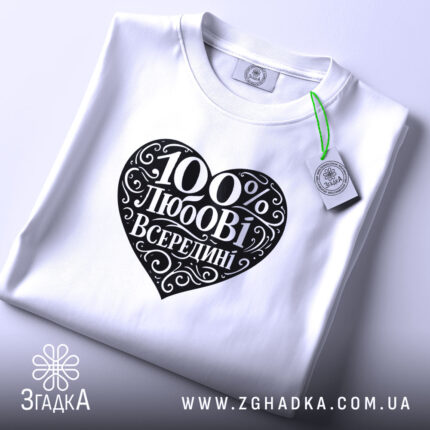 Футболка 100% любові всередині, біла, складена на поверхні, ракурс зверху, фон світло-фіолетовий, принт у формі серця чорного кольору з написом "100% любові всередині" в декоративному шрифті, з етикеткою зеленого кольору.