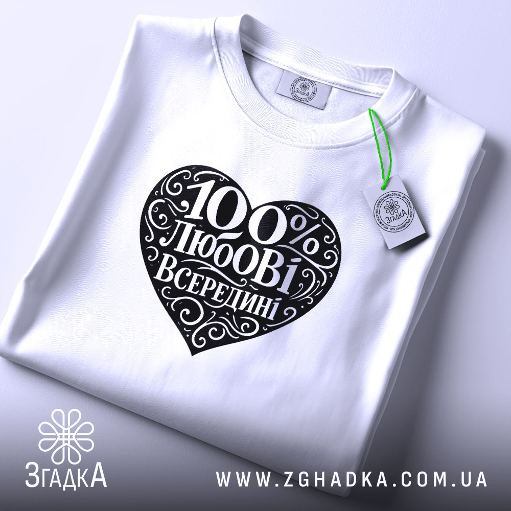 Футболка 100% любові всередині, біла, складена на поверхні, ракурс зверху, фон світло-фіолетовий, принт у формі серця чорного кольору з написом "100% любові всередині" в декоративному шрифті, з етикеткою зеленого кольору.
