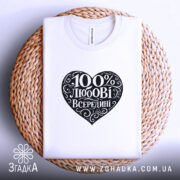Футболка 100% любові всередині, біла, складена на плетеній кошику, ракурс зверху, фон світлий, принт у формі серця чорного кольору з написом "100% любові всередині" в декоративному шрифті.