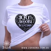 Футболка 100% любові всередині, біла, складена на поверхні, ракурс зверху, фон світло-фіолетовий, принт у формі серця чорного кольору з написом "100% любові всередині" в декоративному шрифті, з етикеткою зеленого кольору.
