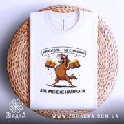 Футболка алкоголь це страшно, біла футболка, що лежить на плетеній основі, з принтом коня, який тримає пивні кухлі, фон світлий, текст на футболці: 'Алкоголь — це страшно. Але мене не налякати.'