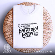 Футболка багатою буду, білого кольору, складена на плетеній кошику, фон світло-сірий, принт чорного кольору з текстом та малюнком дзеркала, видно округлий виріз горловини.