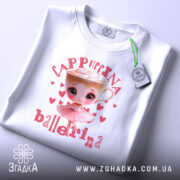 Футболка Ballerina Cappuccina білого кольору, складена на поверхні. Принт з малюнком дівчинки-балерини та чашки капучино з вершками, оточений сердечками, добре видно. На футболці прикріплений етикет з логотипом бренду.