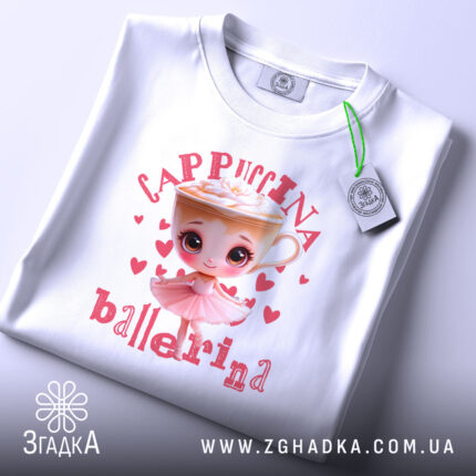 Футболка Ballerina Cappuccina білого кольору, складена на поверхні. Принт з малюнком дівчинки-балерини та чашки капучино з вершками, оточений сердечками, добре видно. На футболці прикріплений етикет з логотипом бренду.