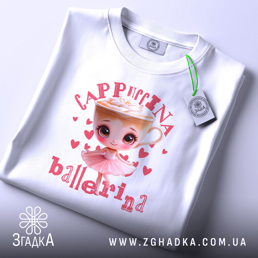 Футболка Ballerina Cappuccina білого кольору, складена на поверхні. Принт з малюнком дівчинки-балерини та чашки капучино з вершками, оточений сердечками, добре видно. На футболці прикріплений етикет з логотипом бренду.