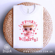 Футболка Ballerina Cappuccina білого кольору, складена на плетеній кошику. Принт з малюнком дівчинки-балерини та чашки капучино з вершками, оточений сердечками, добре видно. Кошик має натуральний колір, що контрастує з футболкою.