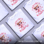 Футболка Ballerina Cappuccina білого кольору, зображена на моделі, яка позує з легким усмішкою. На фоні ніжно-фіолетовий колір, принт з малюнком дівчинки-балерини та чашки капучино з вершками, оточений сердечками. Текст на футболці виконаний у рожевих та червоних тонах.