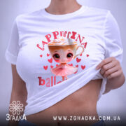 Футболка Ballerina Cappuccina білого кольору, складена на поверхні. Принт з малюнком дівчинки-балерини та чашки капучино з вершками, оточений сердечками, добре видно. На футболці прикріплений етикет з логотипом бренду.