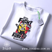 Футболка Бій з кунг-фу, біла, на столі, з верхнього ракурсу, фон світло-фіолетовий, принт з жовтим персонажем у чорному одязі, текстом 'LET'S FIGHT WITH KUNGFU SOUNDS!!!' на червоному фоні, з етикеткою.