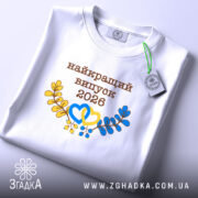 Футболка біла випуск 2026, на столі, з прямим ракурсом, фон світло-сірий, принт з написом 'найкращий випуск 2026' у коричневому кольорі, жовті та блакитні елементи у вигляді сердець і гілок, на комірці є етикетка.