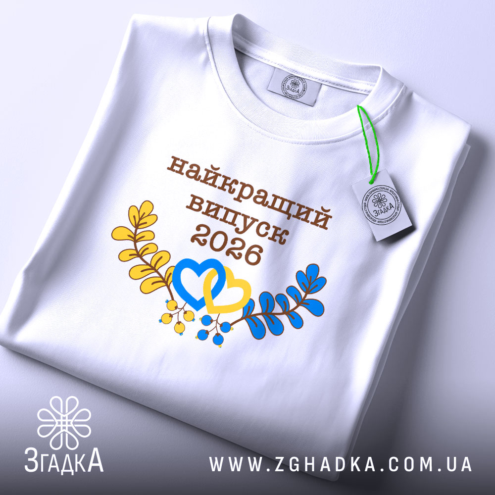 Футболка біла випуск 2026, на столі, з прямим ракурсом, фон світло-сірий, принт з написом 'найкращий випуск 2026' у коричневому кольорі, жовті та блакитні елементи у вигляді сердець і гілок, на комірці є етикетка.