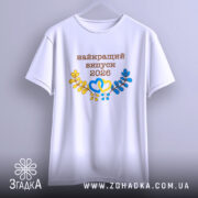 Футболка біла випуск 2026, підвішена на вішалці, фон світло-сірий, принт з написом 'найкращий випуск 2026' у коричневому кольорі, жовті та блакитні елементи у вигляді сердець і гілок, короткі рукави.