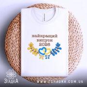 Футболка біла випуск 2026, на плетеній основі, з прямим ракурсом, фон світло-сірий, принт з написом 'найкращий випуск 2026' у коричневому кольорі, жовті та блакитні елементи у вигляді сердець і гілок, видима етикетка на комірці.