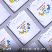 Футболка біла випуск 2026, на моделі, з коротким рукавом, фон світло-сірий, принт з написом 'найкращий випуск 2026' у коричневому кольорі, з жовтими та блакитними елементами у вигляді сердець і гілок.