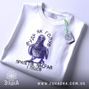 Футболка будь як голуб, біла, на плоскій поверхні, з принтом голуба та текстом, фон світло-сірий, деталі принта чіткі, присутня етикетка з зеленим шнурком.