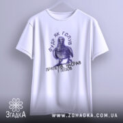 Футболка будь як голуб, біла, підвішена на вішалці, з принтом голуба та текстом, фон світло-сірий, деталі принта чіткі, акцент на формі футболки.