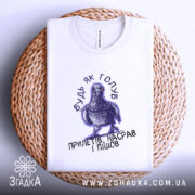 Футболка будь як голуб, біла, на плоскій поверхні, з принтом голуба та текстом, фон світло-сірий, деталі принта чіткі, розташована на плетеній основі.