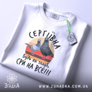 Футболка будь як голуб сри на все. Біла футболка, розкладена на поверхні, з видимим принтом, що містить двох голубів на кольоровому фоні. Принт супроводжується написом 'СЕРГІВНА, будь як голуб, сри на все!!!'. Фон світло-фіолетовий, з яскравим зеленим ярликом.