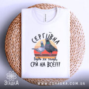 Футболка будь як голуб сри на все. Біла футболка, складена на плетеній кошику, з принтом, що містить двох голубів на кольоровому фоні. Напис 'СЕРГІВНА, будь як голуб, сри на все!!!' розташований під зображенням. Фон світло-фіолетовий.