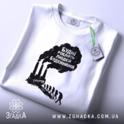 Футболка будні роблять людей буденними. Біла футболка, складена на поверхні, фон світло-сірий. Принт чорного кольору з текстом та зображенням димарів і силуетів людей, що йдуть, з зеленим ярликом.