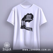 Футболка будні роблять людей буденними. Біла футболка, підвішена на вішалці, фон світло-сірий. Принт чорного кольору з текстом та зображенням димарів і силуетів людей, що йдуть.