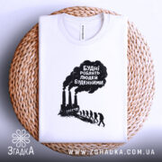 Футболка будні роблять людей буденними. Біла футболка, складена на плетеній кошику, фон світло-сірий. Принт чорного кольору з текстом та зображенням димарів і силуетів людей, що йдуть.