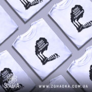 Футболка будні роблять людей буденними. Біла футболка з коротким рукавом, зображена на молодому чоловікові, фон світло-фіолетовий. Принт чорного кольору з текстом та зображенням димарів і силуетів людей, що йдуть.