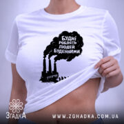 Футболка будні роблять людей буденними. Біла футболка, складена на поверхні, фон світло-сірий. Принт чорного кольору з текстом та зображенням димарів і силуетів людей, що йдуть, з зеленим ярликом.