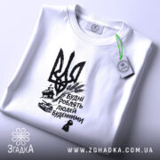 Футболка будні роблять людей буденними. Біла футболка, зображена на плоскій поверхні, фон світло-сірий. Принт чорного кольору з тризубом та написом 'Будні роблять людей буденними'. На футболці видно етикетку з зеленим шнурком.