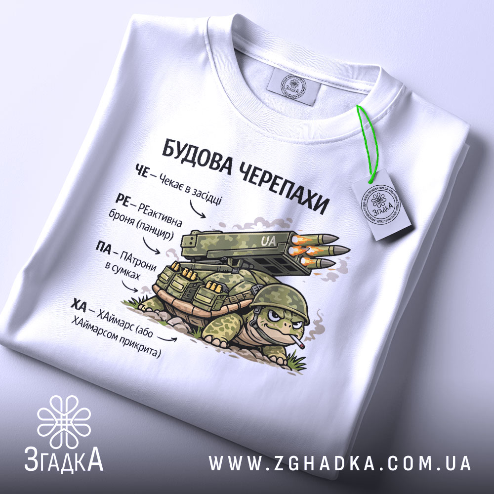 Футболка будова черепахи, біла, зображена зверху, фон світло-сірий, принт з черепахою та ракетною установкою, текст на українській мові, на комірці видна етикетка.
