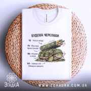 Футболка будова черепахи, біла, складена на плетеній основі, фон нейтральний, принт з черепахою та ракетною установкою, текст на українській мові, видно деталі принта.