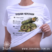Футболка будова черепахи, біла, зображена зверху, фон світло-сірий, принт з черепахою та ракетною установкою, текст на українській мові, на комірці видна етикетка.