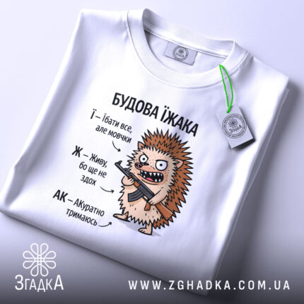 Футболка будова їжака, біла, на столі, ракурс зверху, фон світло-сірий, принт з їжаком, який тримає гвинтівку, текст з описом характеристик їжака, на етикетці зелена стрічка.