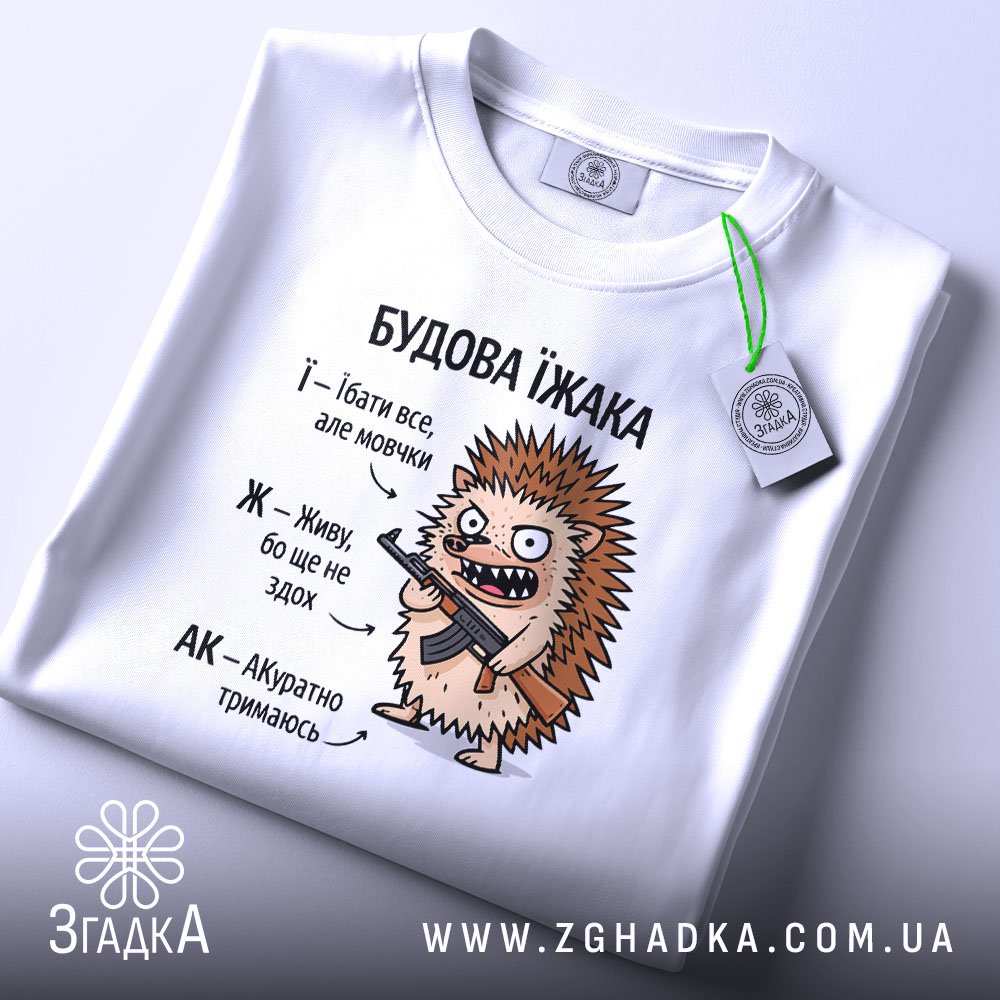 Футболка будова їжака, біла, на столі, ракурс зверху, фон світло-сірий, принт з їжаком, який тримає гвинтівку, текст з описом характеристик їжака, на етикетці зелена стрічка.