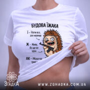 Футболка будова їжака, біла, на столі, ракурс зверху, фон світло-сірий, принт з їжаком, який тримає гвинтівку, текст з описом характеристик їжака, на етикетці зелена стрічка.