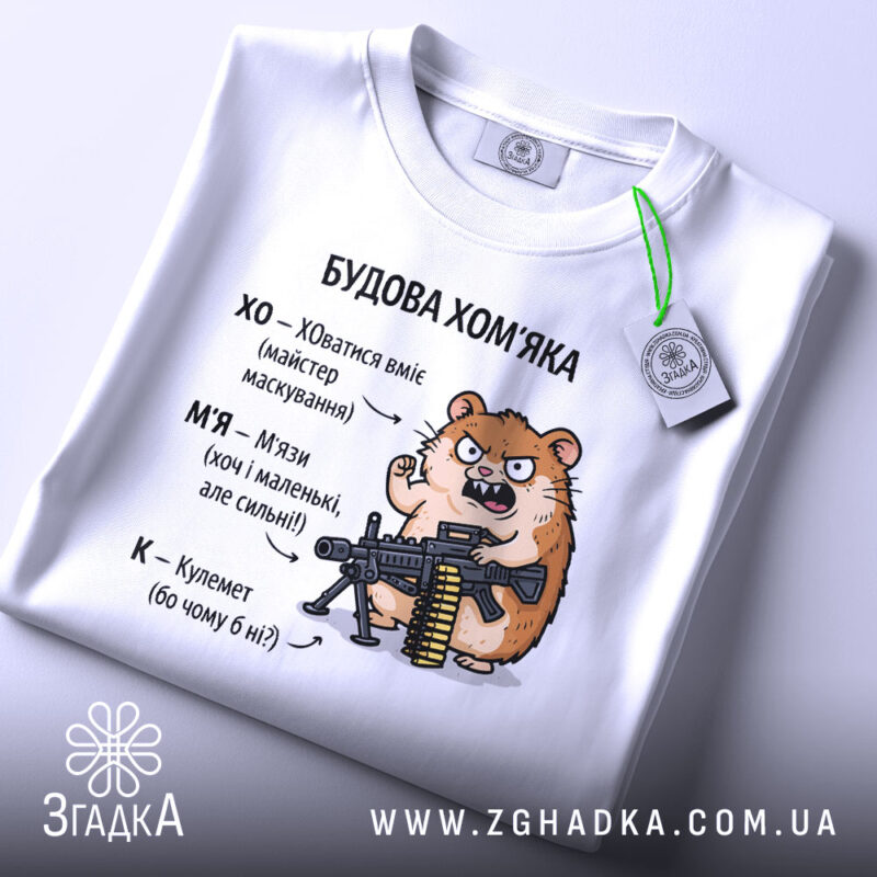 Футболка будова хомʼяка, біла, з верхнього ракурсу, фон світлий, принт з хомʼяком, що тримає гвинтівку, текст з характеристиками хомʼяка, на ярлику зелена стрічка.