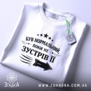 Футболка був нормальний поки не зустрів її, біла футболка, складена на поверхні, фон світло-сірий, принт чорного кольору з текстом та стрілкою, на комірці видна етикетка.
